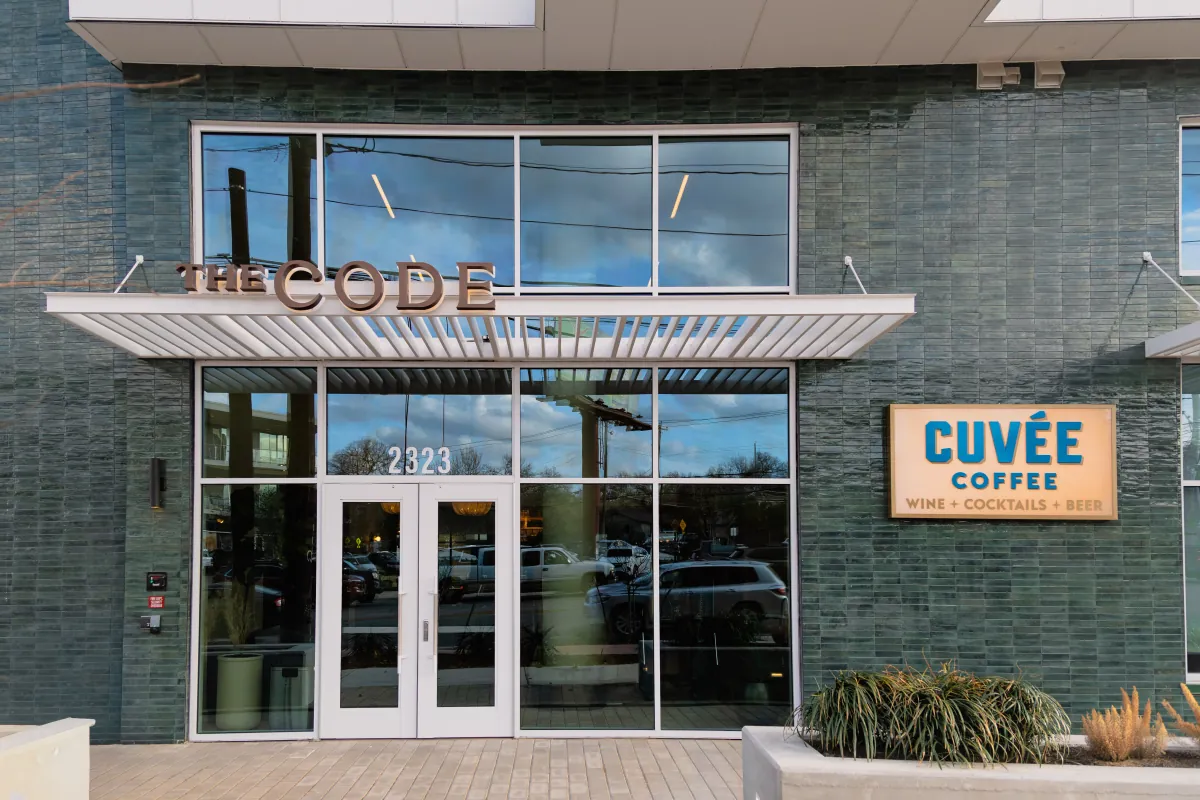 The glass front of The Code in South Austin, with a Cuvée Coffee sign on the dark brick wall beside the doors
