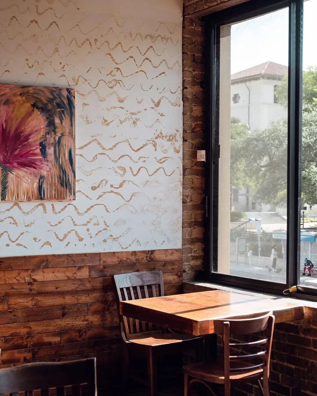A wooden table and chairs by the window of the Medici café on Guadalupe, with a painting on a white relief wall