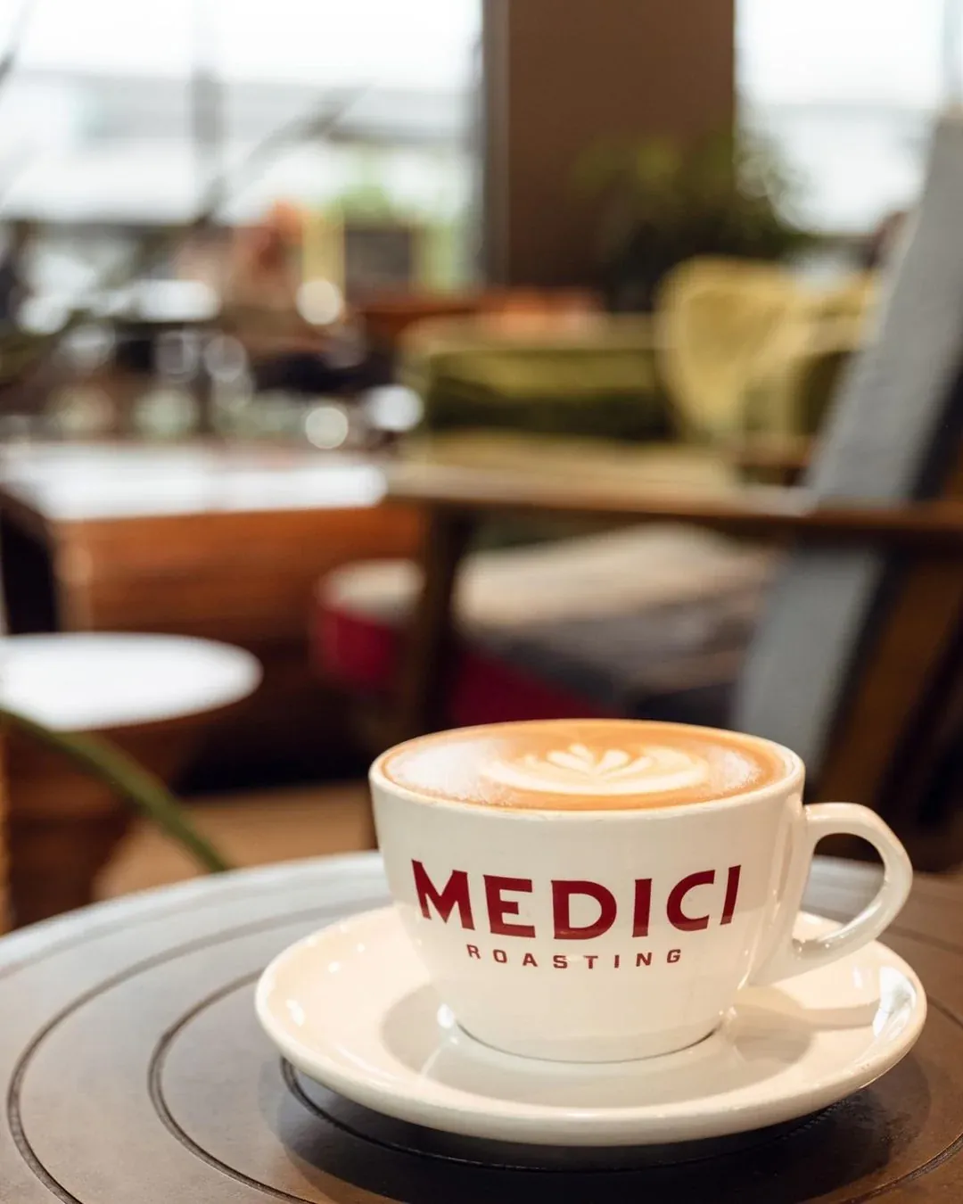 A cappuccino in a white Medici Roasting cup on a wooden table in the Springdale General café, with armchairs and tables behind