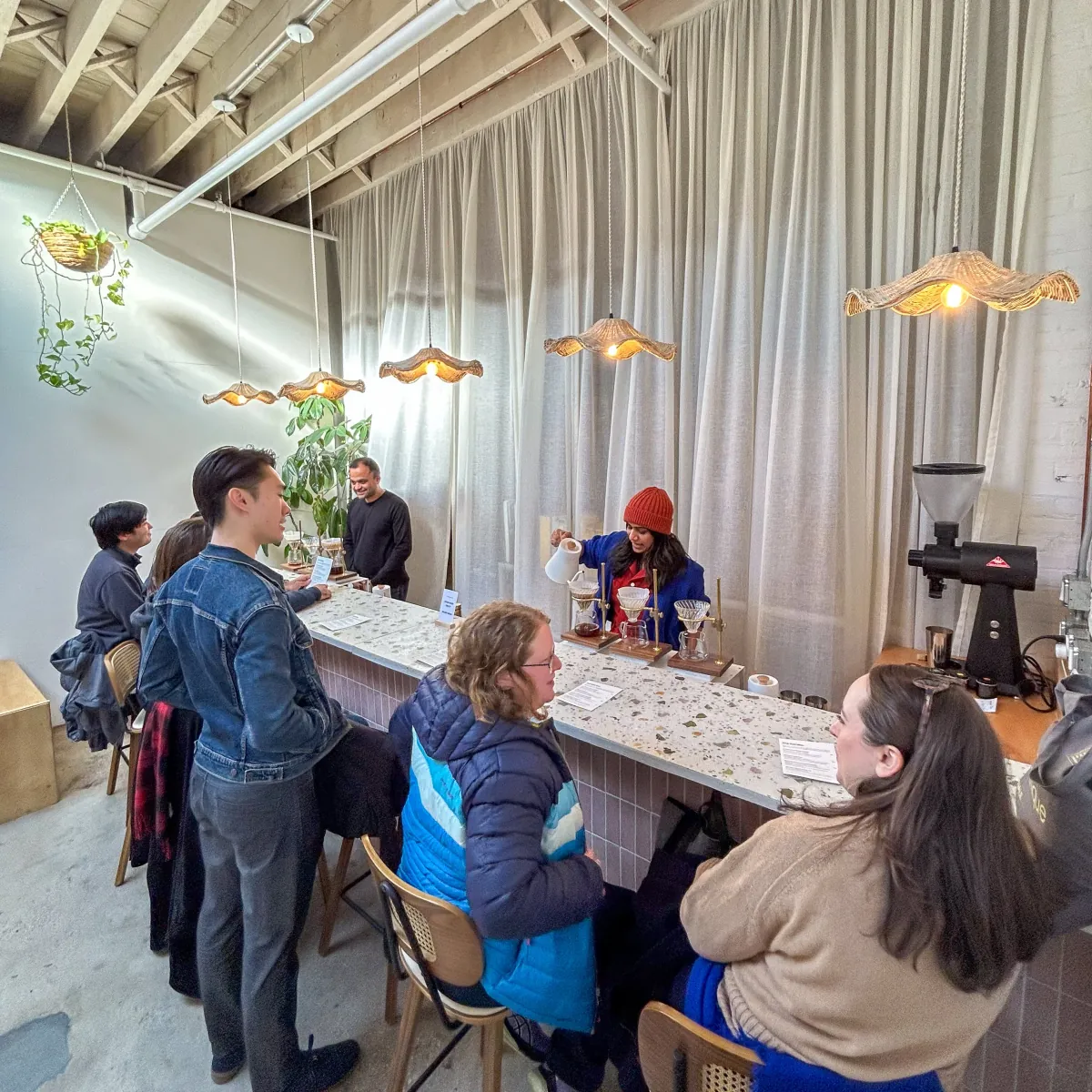 Guests on stools at the terrazzo pour-over bar in the Driftaway tasting room while a barista pours water over three V60 brewers under rattan lampshades