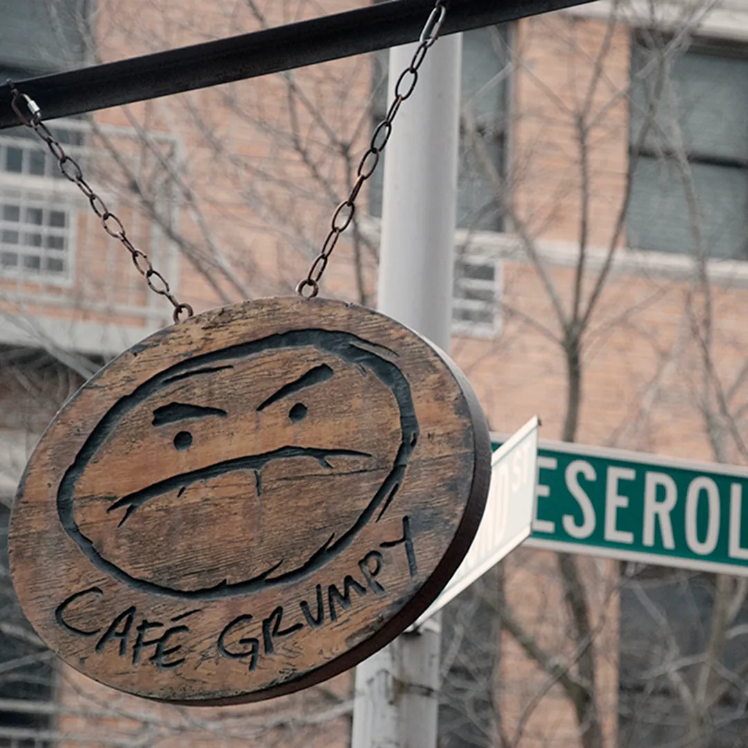 The carved wooden Café Grumpy sign with its frowning face hanging from chains beside the Meserole Avenue street sign
