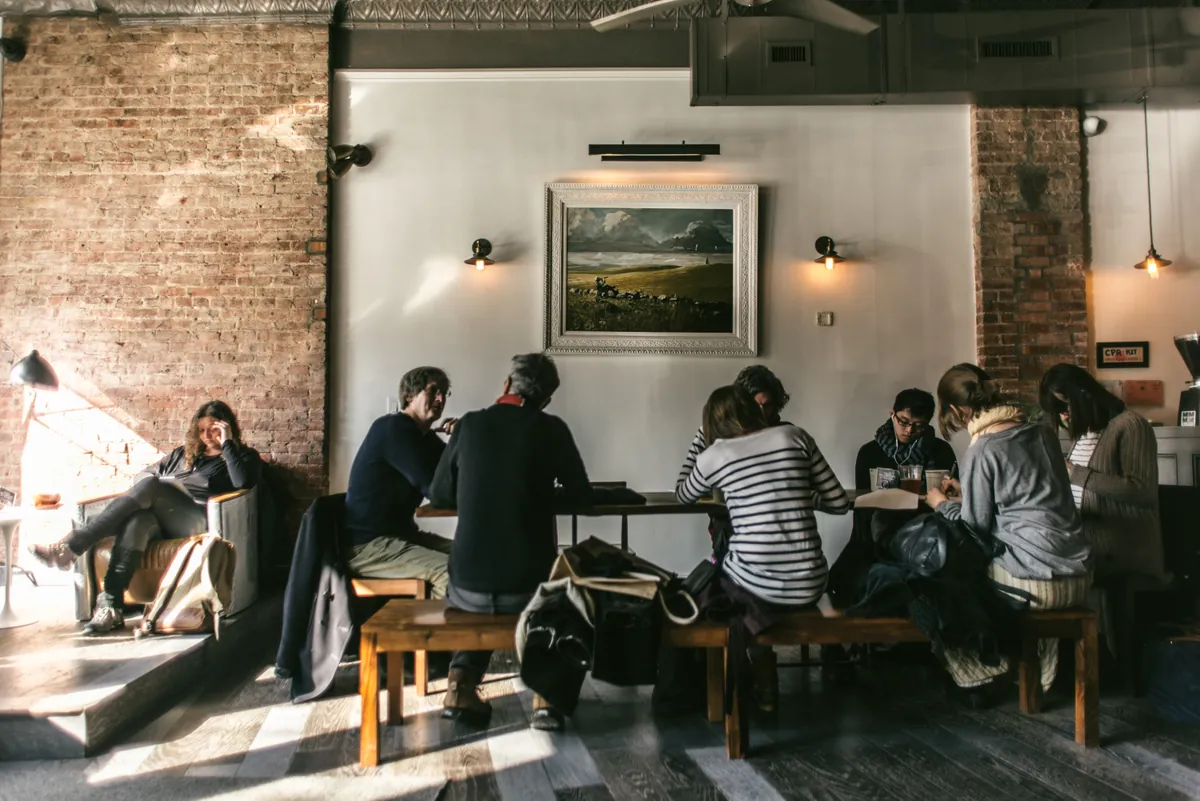 People on wooden benches at a long table under a framed landscape painting between exposed brick walls at Hungry Ghost in Prospect Heights