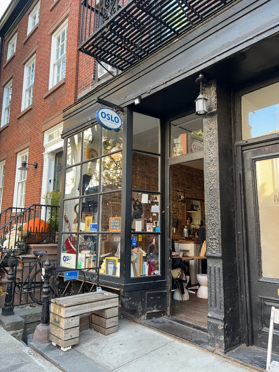 The black-framed shopfront of Oslo Coffee in the West Village, with the blue Oslo sign in the window, a wooden bench outside and customers at the counter inside