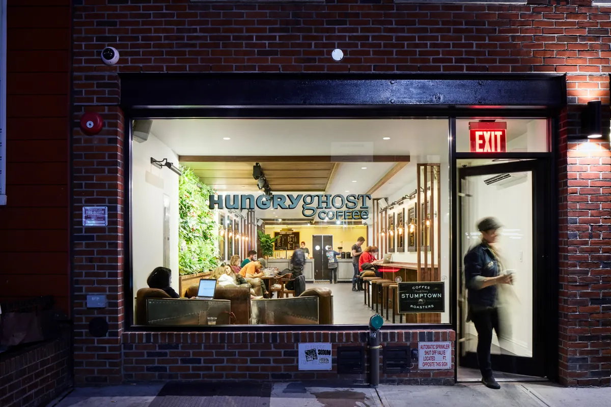 The Hungry Ghost Coffee shopfront on Metropolitan Avenue at night, its big window showing leather armchairs, a living wall and people at the counter under the shop's lettering