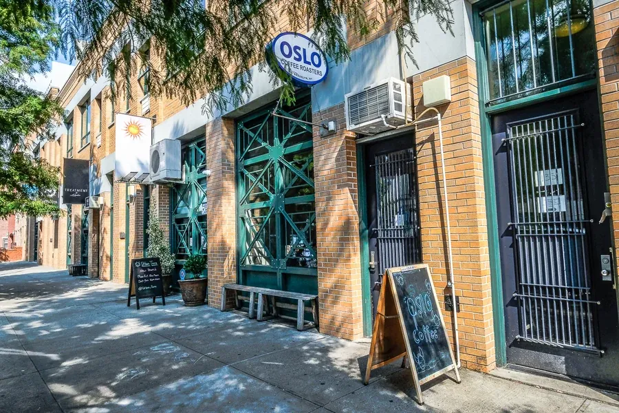 The Oslo Coffee Roasters shop on Roebling Street in Williamsburg, with a blue oval sign over green-framed windows, a wooden bench and a chalkboard on the pavement under a tree