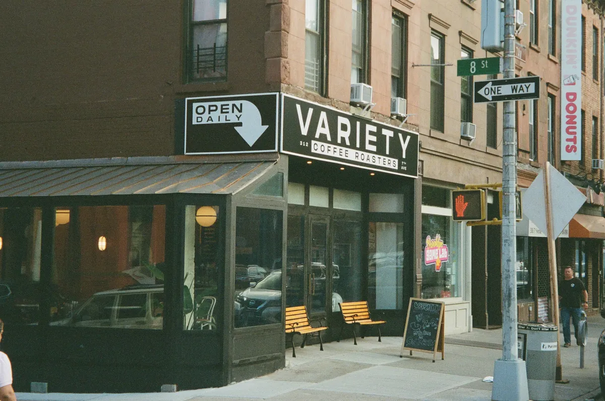 The corner shopfront of Variety Coffee Roasters in Park Slope, with a black Variety sign over the door, two wooden benches and a chalkboard on the pavement