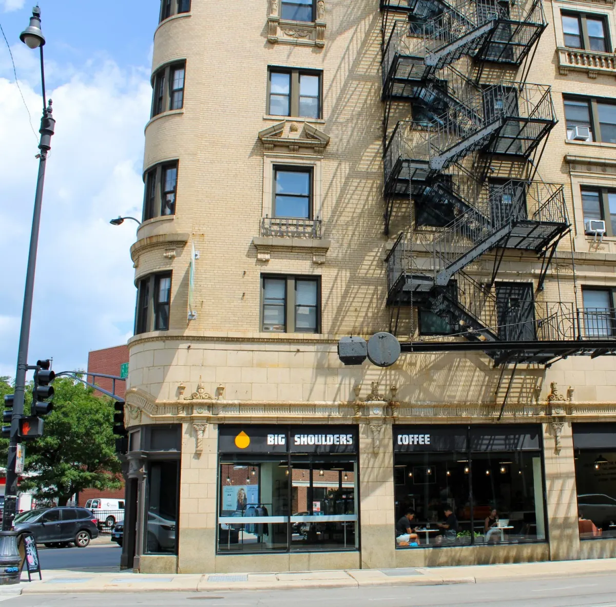 The Big Shoulders Coffee café on the ground floor of a pale brick corner building in Lincoln Park, with fire escapes above and a black awning over the windows