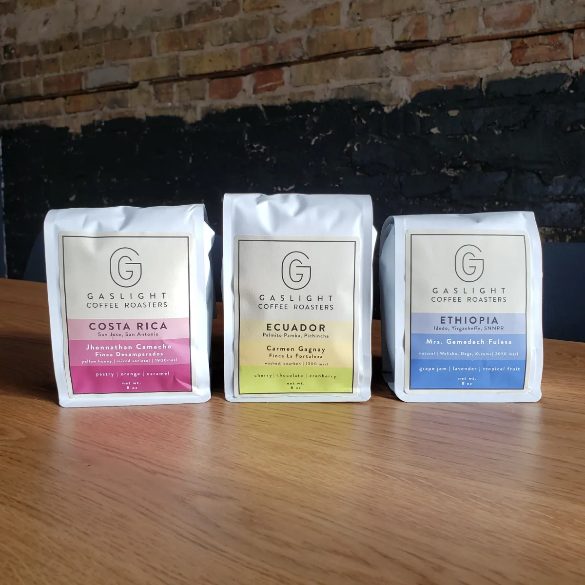Three white Gaslight Coffee Roasters bags, Costa Rica, Ecuador and Ethiopia, lined up on a wooden table in front of a brick wall