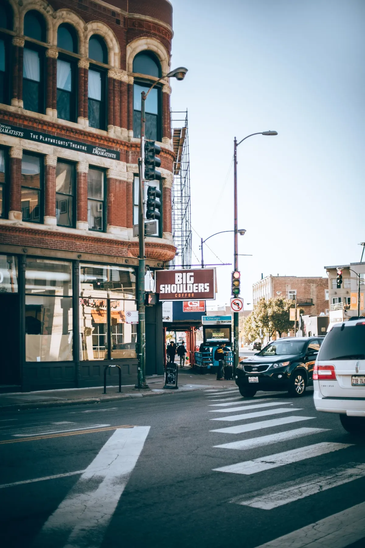 The Big Shoulders Coffee café in the ground floor of a red-brick corner building in West Town, seen across a street crossing with a car waiting at the lights