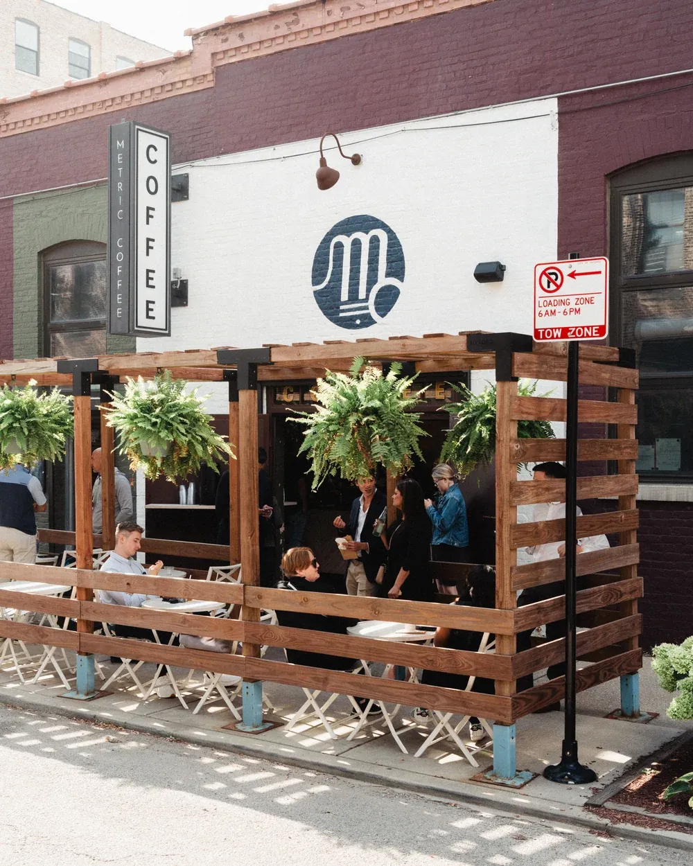 The wooden street patio of the Metric café in the Kinzie Corridor, with hanging ferns, folding chairs and the white M sign over the door