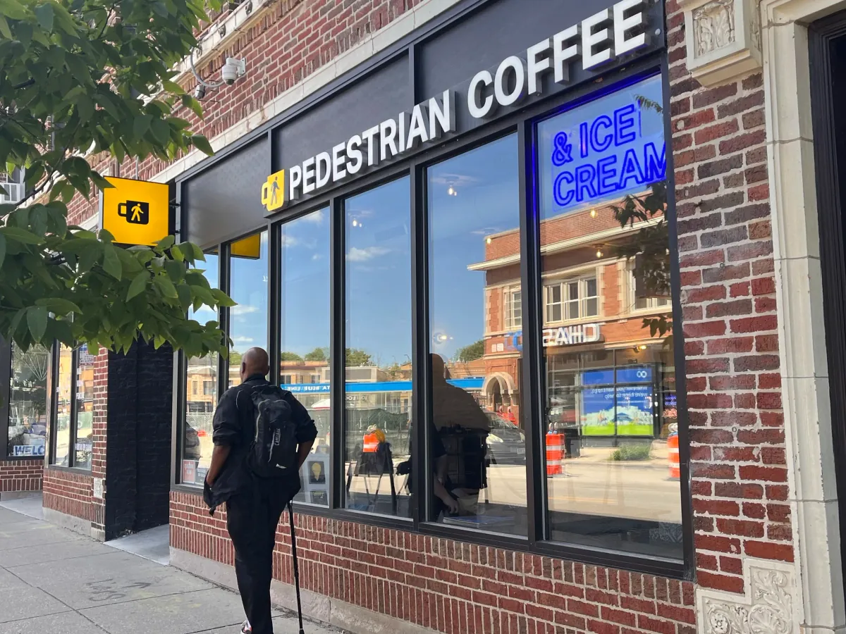 The Pedestrian Coffee and Ice Cream shopfront in Logan Square, a brick corner with a lit sign and a blue neon ice cream sign in the window, and a man walking past under a tree