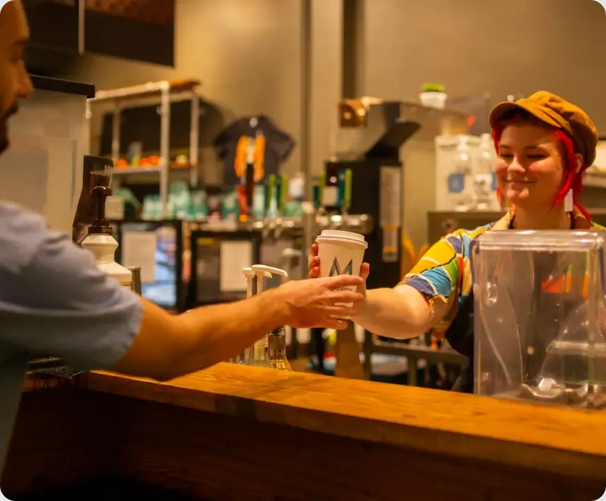 A barista in a flat cap hands a paper cup of coffee across the wooden counter of the Metropolis Edgewater Café
