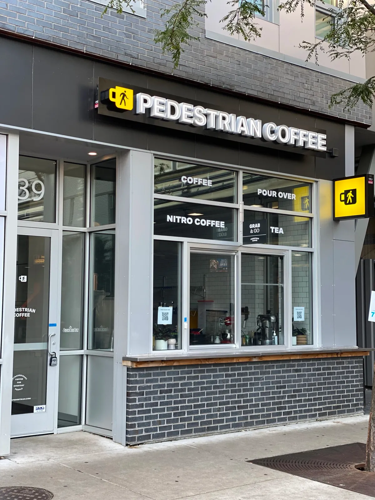 The black fascia and lit Pedestrian Coffee sign over the Belmont café, with coffee, pour over, nitro coffee, grab and go and tea lettered on the windows above a brick sill