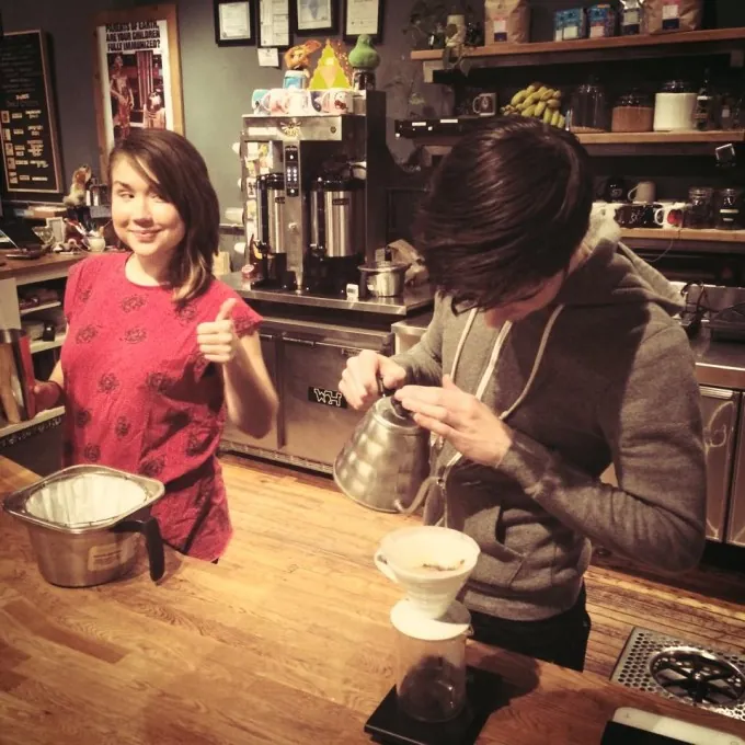 Two baristas behind the wooden bar at The Wormhole, one pouring a pour-over from a gooseneck kettle, with batch brewers and shelves of mugs and bags behind