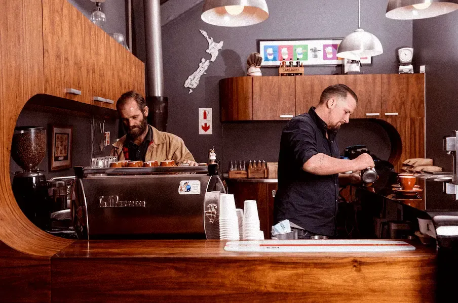 Two baristas at work behind the curved wooden bar of a Deluxe Coffeeworks café, a La Marzocco machine between them and a map of New Zealand on the wall