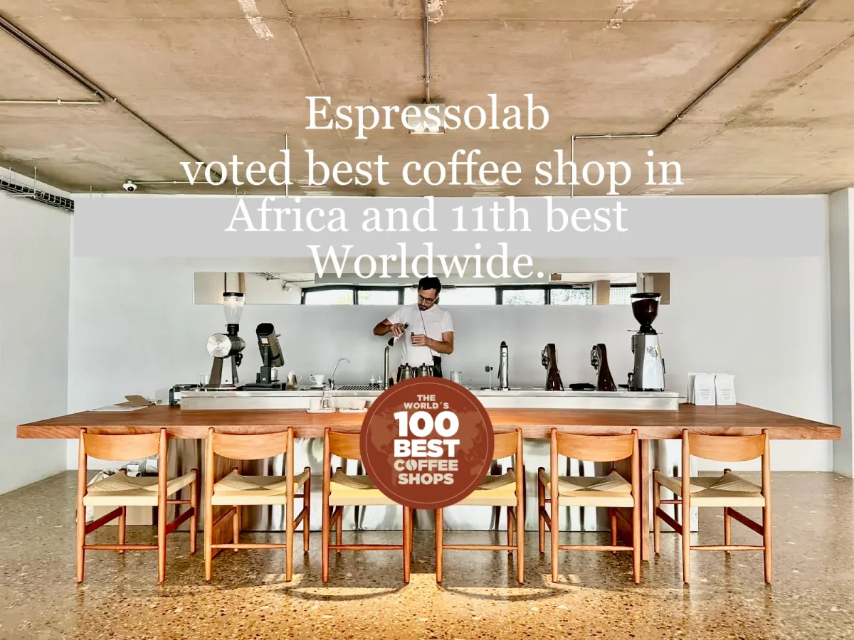 A barista behind the long wooden communal table and brew bar of Espressolab's flagship store, with grinders and brewers lined up under a concrete ceiling