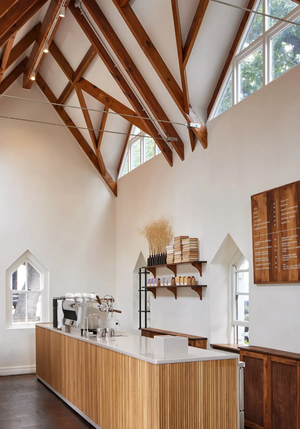 The bar of a Pauline’s café under a pitched timber-trussed ceiling, with a chrome espresso machine on a slatted wooden counter, bags of coffee on shelves and a wooden menu board