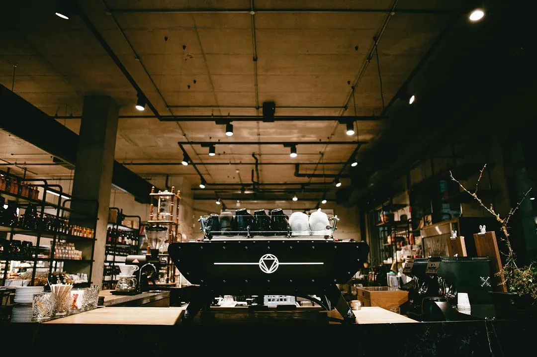 The bar of a Rosetta Roastery café seen from the counter, a black La Marzocco espresso machine in the centre under a concrete ceiling, with shelves of retail goods and a cold-drip tower behind
