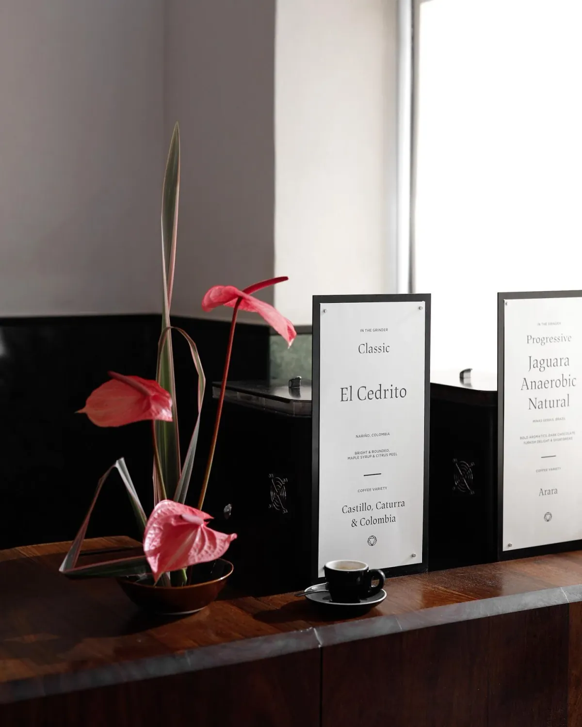 Two grinder cards on the counter of Rosetta Roastery's Bree Street café, one for a Classic Colombian espresso and one for a Progressive Brazilian natural, beside an espresso cup and red anthuriums