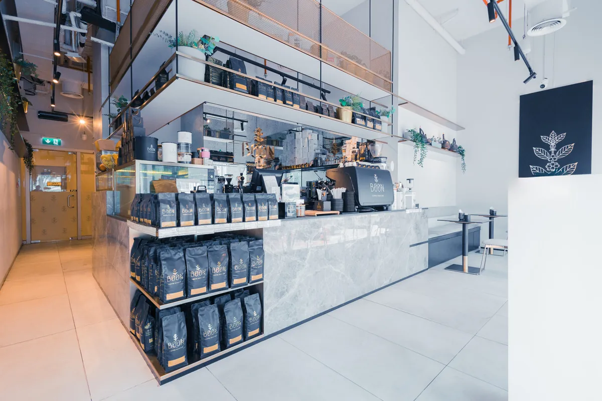 The grey marble counter at Boon Coffee JLT stacked with black bags of beans under floating shelves of coffee, with a black espresso machine and a few tables along the white wall