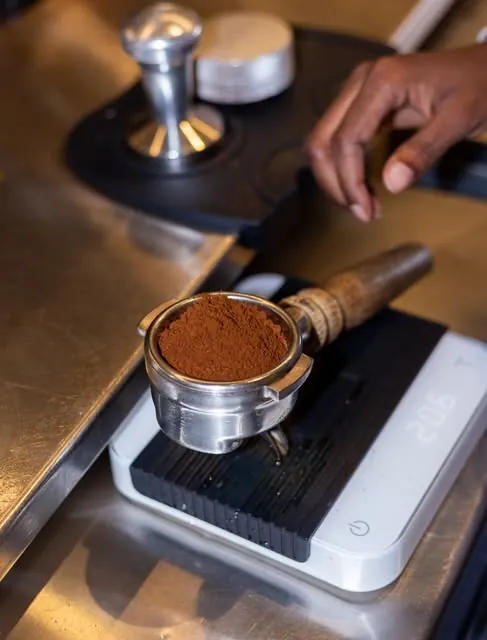 A portafilter heaped with freshly ground coffee resting on a white scale beside a tamper on Drop's espresso bar, with a barista's hand reaching towards it