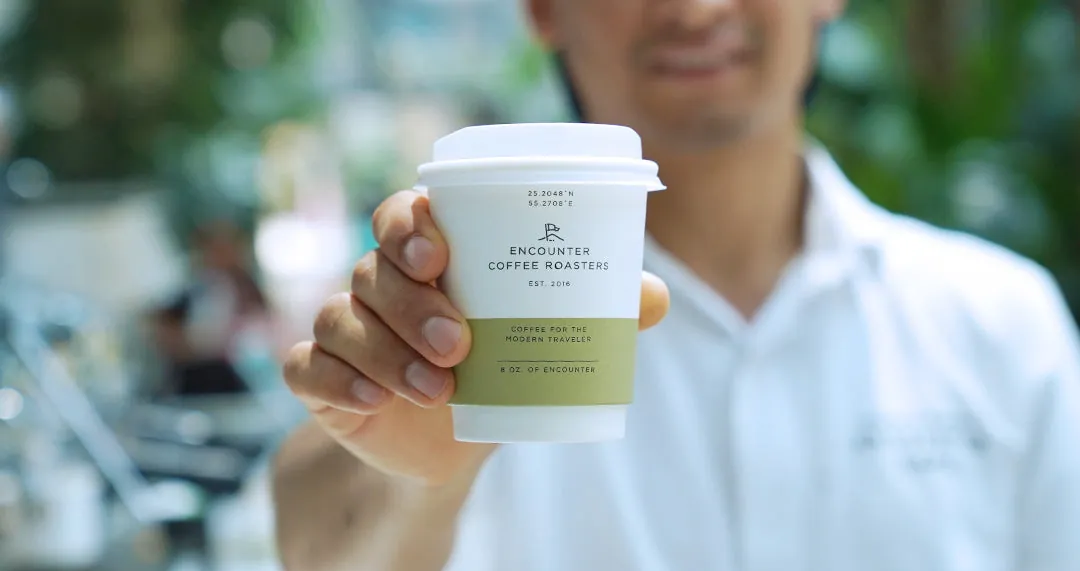 A man in a white shirt holds out a white Encounter Coffee Roasters takeaway cup printed with the line Coffee for the modern traveler