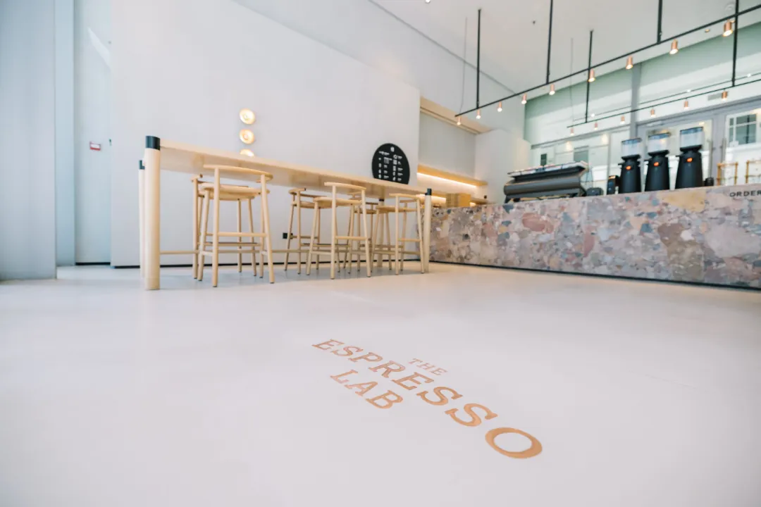 Inside The Espresso Lab's café in Dubai Design District, with the name set in copper letters on a pale floor, a long pale wood table with stools and a counter of pink and grey terrazzo holding grinders and an espresso machine