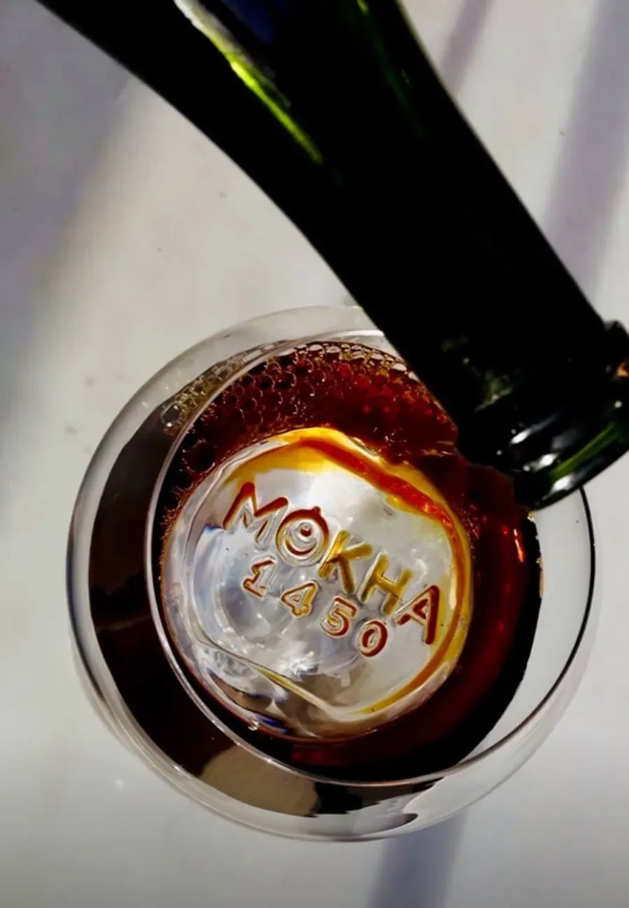 Cold brew poured from a dark green bottle into a double-walled glass stamped Mokha 1450 on its base