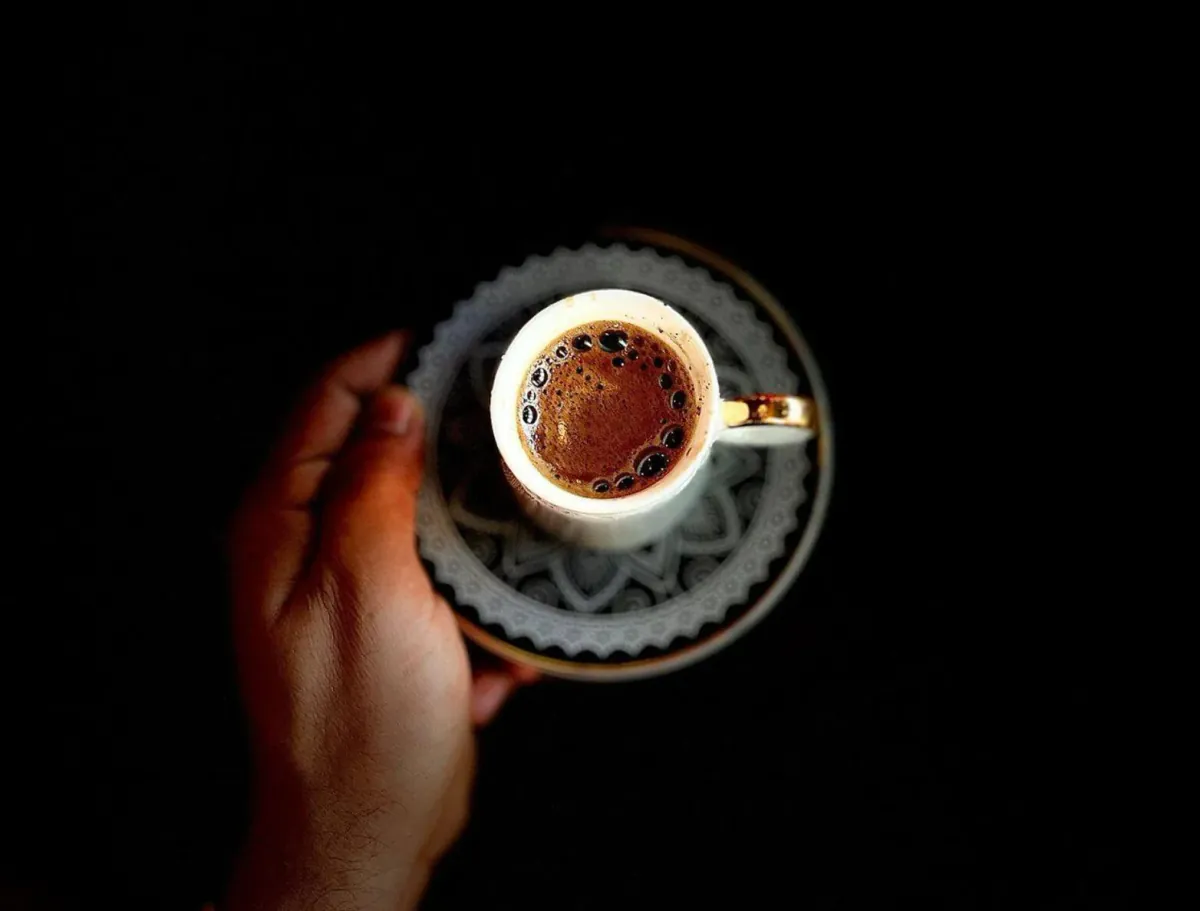 A hand holds a small white cup of black coffee with a gold handle on a patterned saucer against a dark background