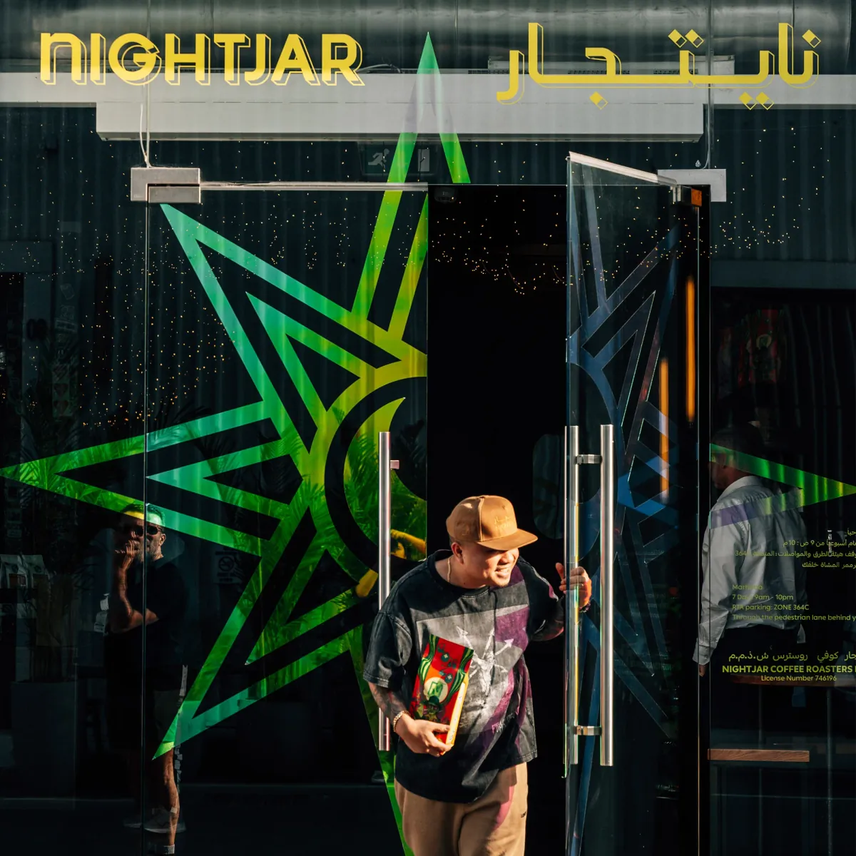The glass doors of Nightjar in Alserkal Avenue, printed with a green star and the name in Latin and Arabic script, as a man in a cap walks out holding a bag of coffee