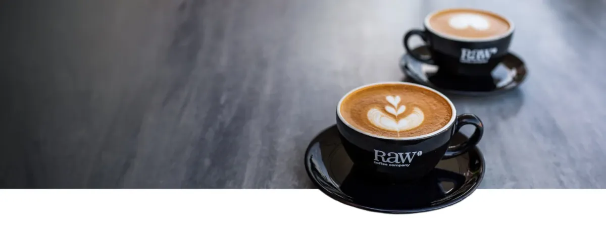 Two flat whites with latte art in black RAW Coffee Company cups and saucers on a grey table