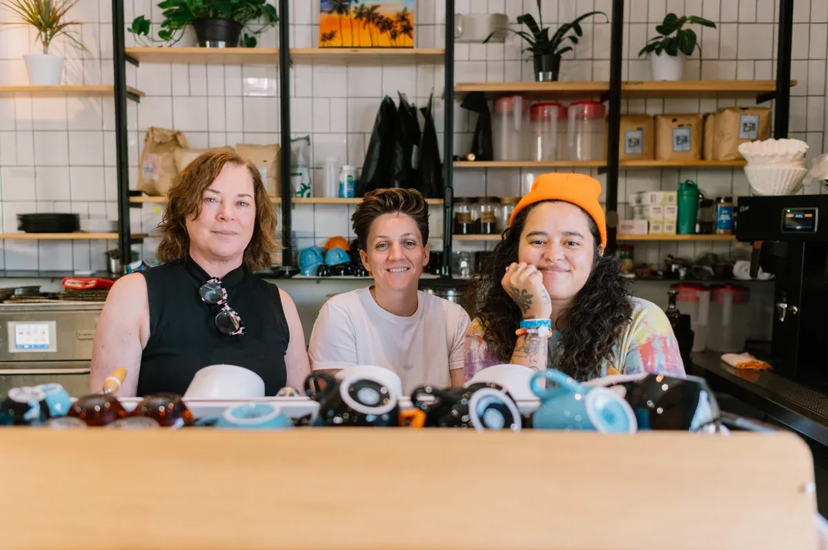 Three members of the Alana's Coffee Roasters team behind the counter of a white-tiled café, with cups and jars on the shelves behind them