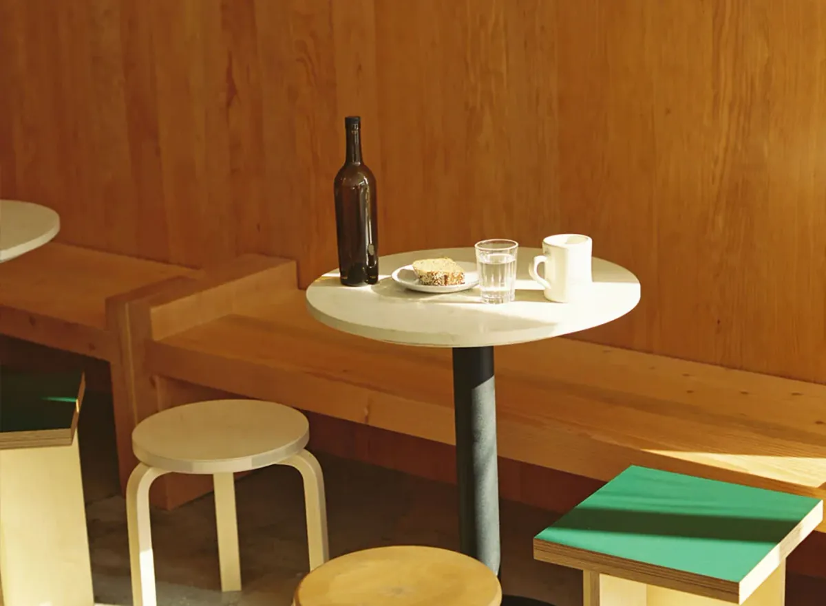 A round white table with a bottle, a glass and a plate between wooden benches and low stools against the wood-panelled wall of Canyon Echo Park