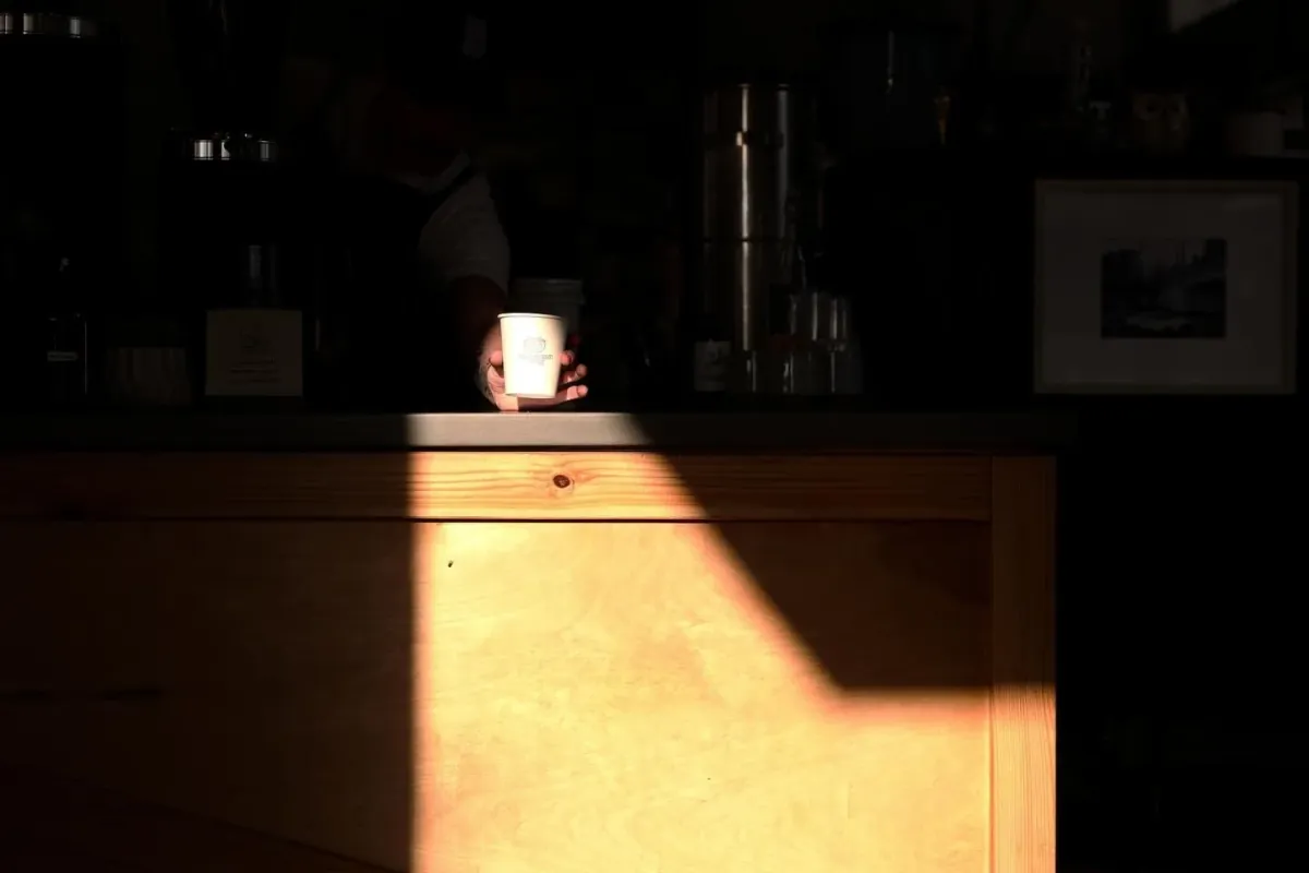 A single cup lit by a shaft of sunlight on the dark counter of Cognoscenti's roastery café, with the espresso machine and shelves in shadow behind it