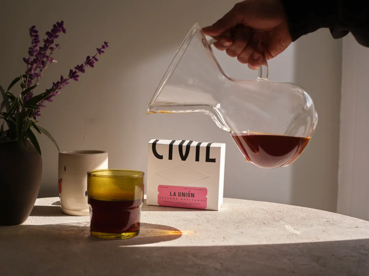 A hand pours coffee from a glass carafe into a tumbler on a stone table beside a pink Civil coffee bag, a mug and a vase of lavender