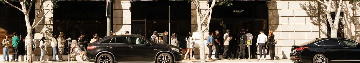 A queue along the stone facade beneath the trees outside the Kumquat DTLA café