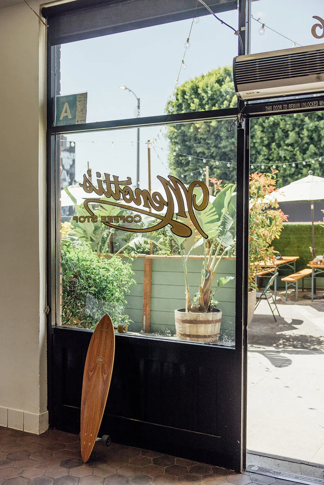 The gold-lettered glass door of Menotti's Venice Beach, with a longboard propped against it and the patio beyond