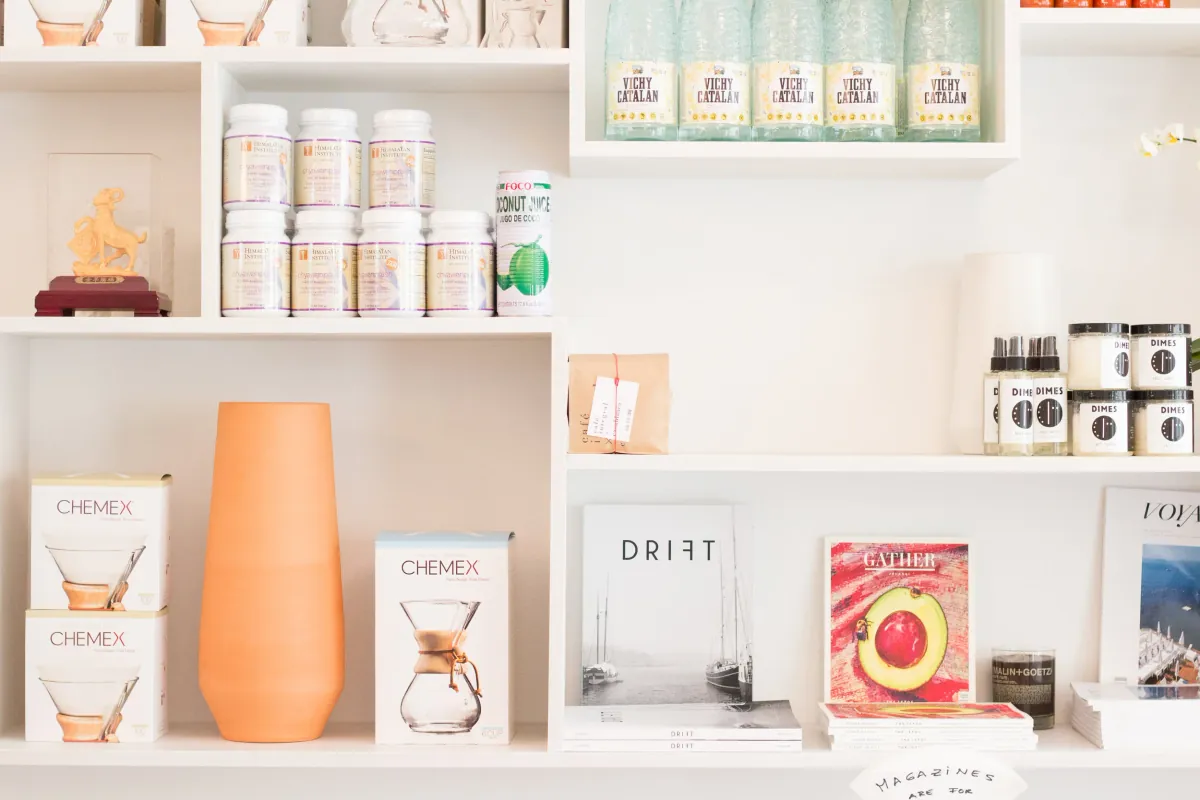 White retail shelves inside Café Integral with Chemex boxes, a terracotta vase, a bag of Café Integral coffee, magazines and bottles of Vichy Catalan water