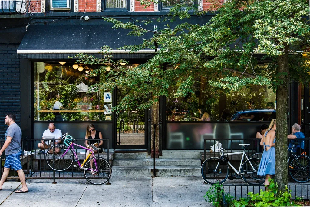 The black shopfront of Ninth Street Espresso in Alphabet City under a tree, with people on the benches outside and two bicycles leaning on the railings