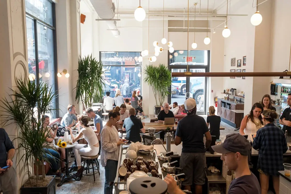 A busy Think Coffee café with a barista at the espresso machine in the foreground, tall windows, potted palms and globe pendant lights over the tables