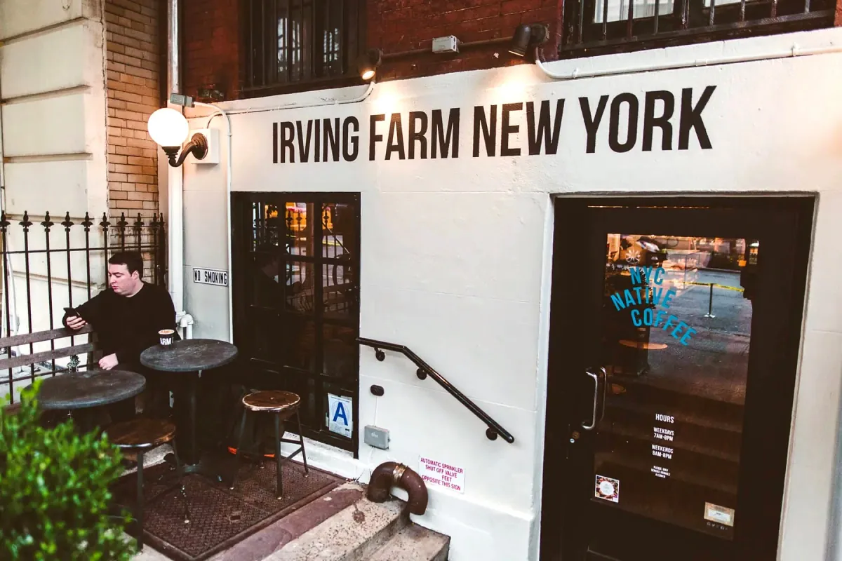 The white-painted front of an Irving Farm New York café below street level, with the name lettered above the door and a man at a small round table outside