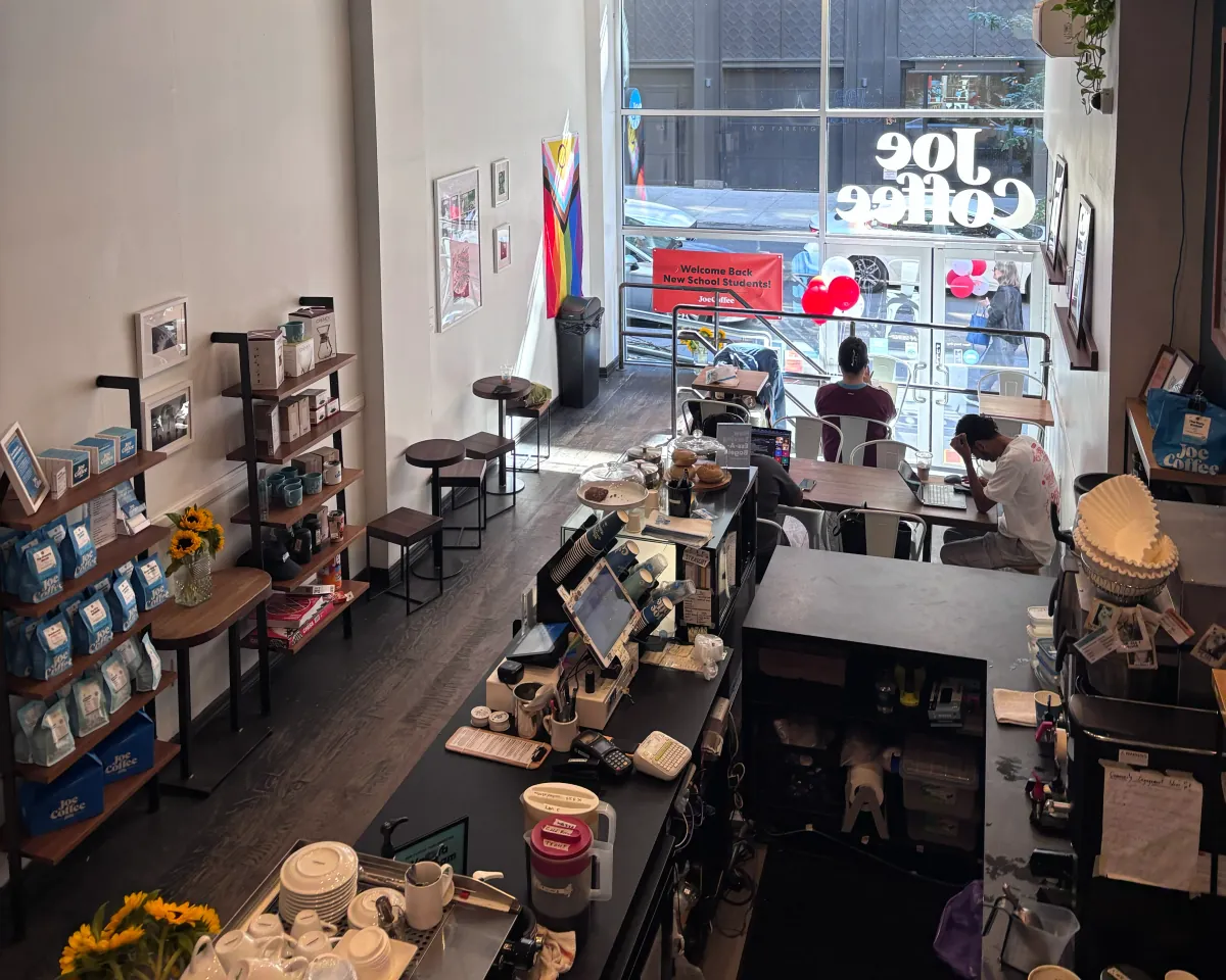 The Joe Coffee café on 13th Street seen from above, with shelves of blue coffee bags along the wall, the counter in the middle and guests at tables by the window