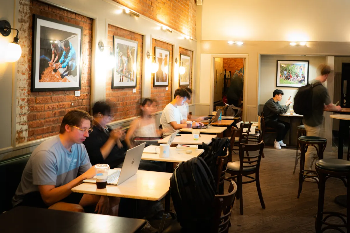 Guests on laptops at small tables inside a Think Coffee café, with framed photographs of coffee producers on the exposed brick wall