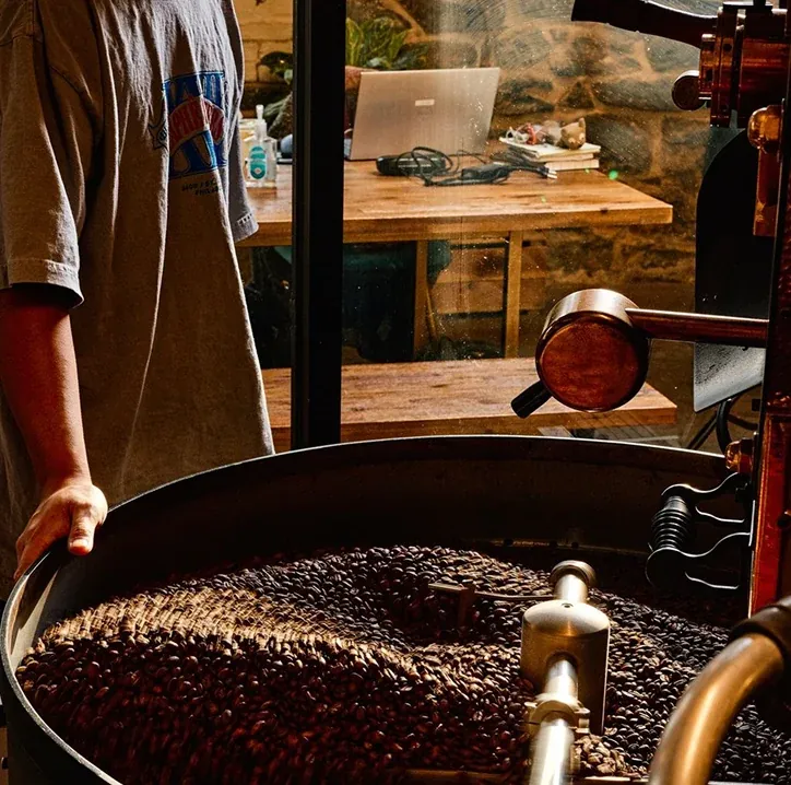 A roaster in a grey T-shirt rests a hand on the cooling tray of the Càphê roaster as the beans turn, with a stone wall and a wooden desk behind