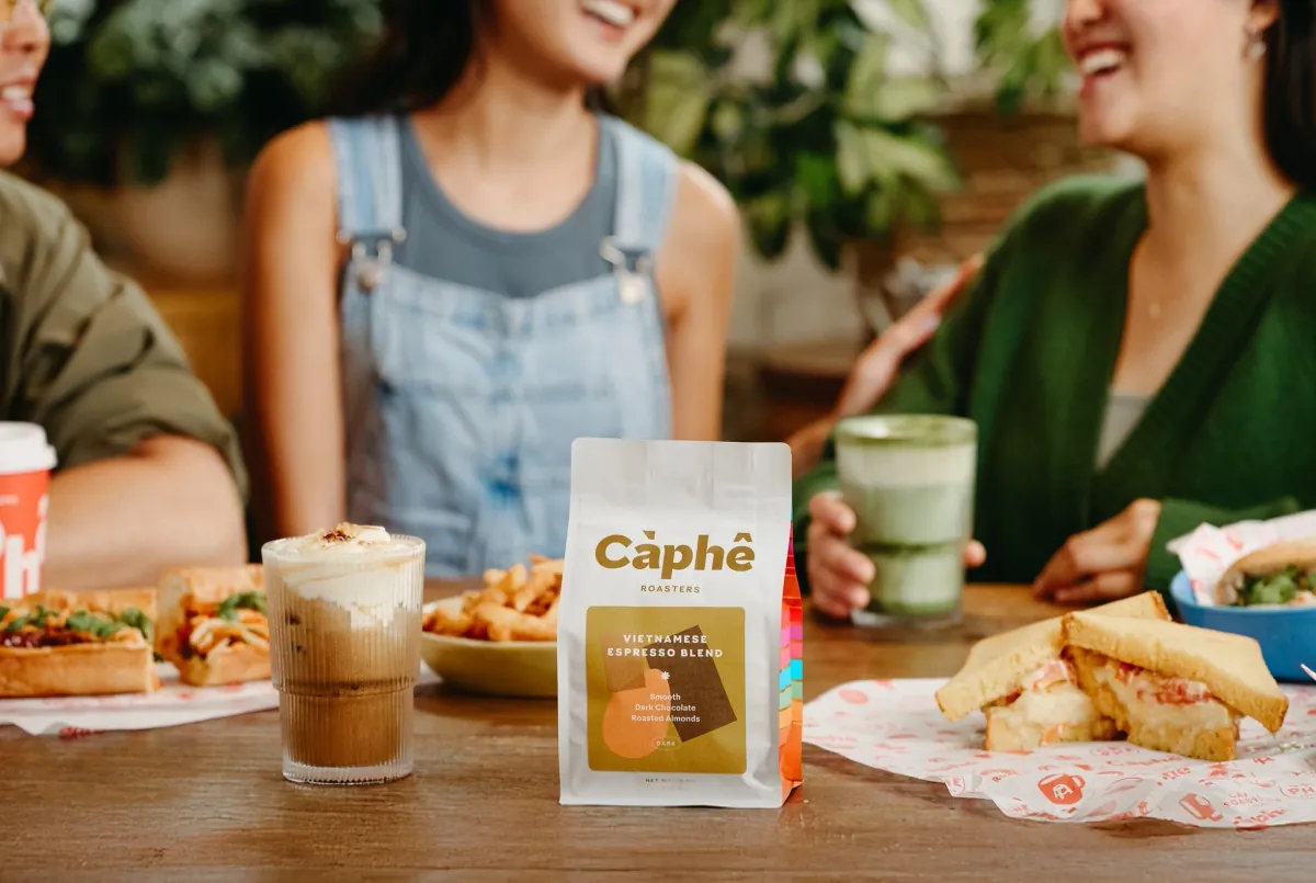A bag of Càphê Roasters Vietnamese Espresso Blend on a wooden table between an iced coffee, a matcha, fries and a bánh mì, with three friends laughing behind