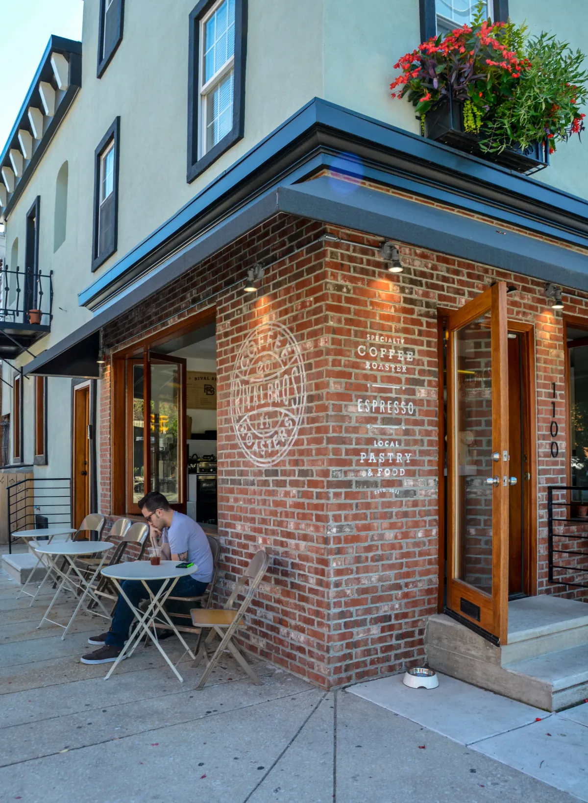 The red-brick corner of the Rival Bros East Passyunk café, painted with the roaster's crest and the words specialty coffee roaster, espresso, local pastry and food, with a man at a folding table outside