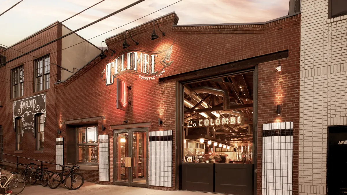 The red-brick La Colombe Fishtown café at dusk, its sign reading La Colombe Torréfaction above a wide open garage door, warm light on the counter inside and bicycles parked by the kerb