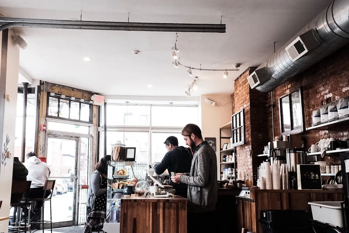 Inside an Ultimo café, a barista at the butcher-block counter with a customer in a grey cardigan, exposed brick and shelves of coffee bags on the right and the glass front door beyond