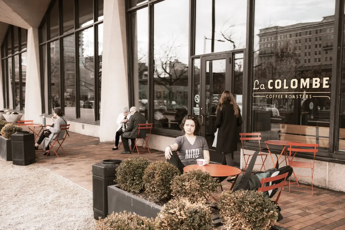 The glass front of the La Colombe Independence Mall café with its gold window lettering, people at red bistro tables on the brick terrace and low box hedges in planters