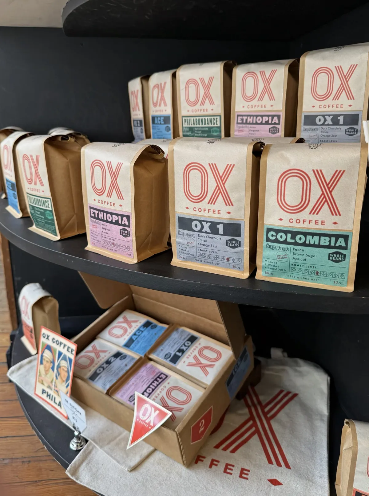 Kraft bags of Ox Coffee lined up on a black shelf, their pastel labels reading Ethiopia, OX 1, Colombia, Ace and Philabundance, with a box of bags and a tote below